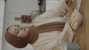 Young woman in hijab working remotely on a tablet computer. The woman eats roasted hazelnuts while she works on the computer in her home kitchen. Freelance concept.Vertical video.