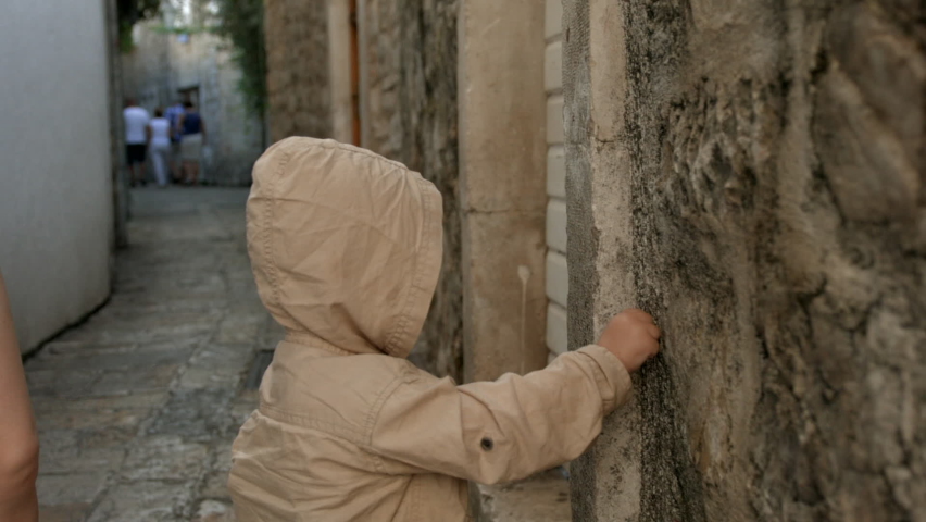 A family walking through the narrow streets. A little boy with his mother who walks around in a brown jacket with a hood and touches brown walls.