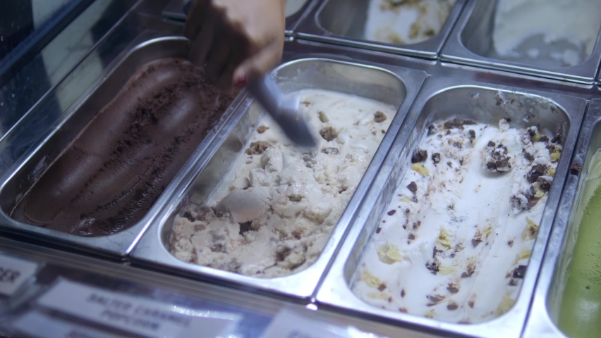 A showcase with different ice cream in containers, a woman scoops ice cream into a waffle cup with a spoon. Consumption of sweets with a high content of sugar and milk fat.
