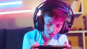 Serious teenager kid with headset playing video game on mobile phone at home - concept of relaxation, technology addiction and entertainment