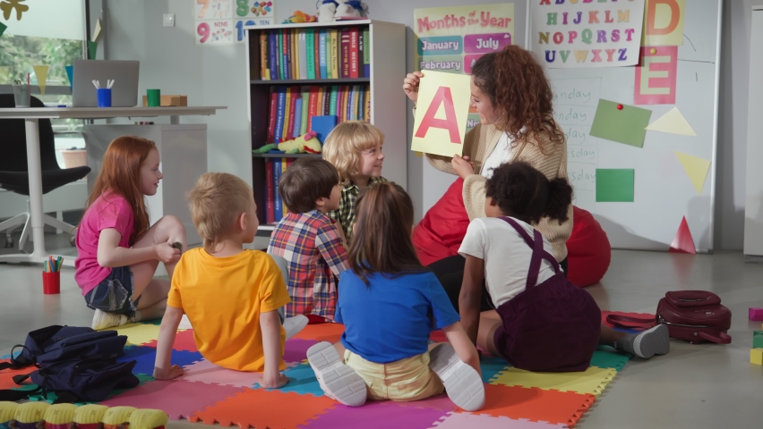 Female teacher hold flashcard with letter a teaching preschool kids alphabet. Diverse pupils of primary school sit on floor with teacher having alphabet lesson Royalty-Free Stock Footage #1093403253