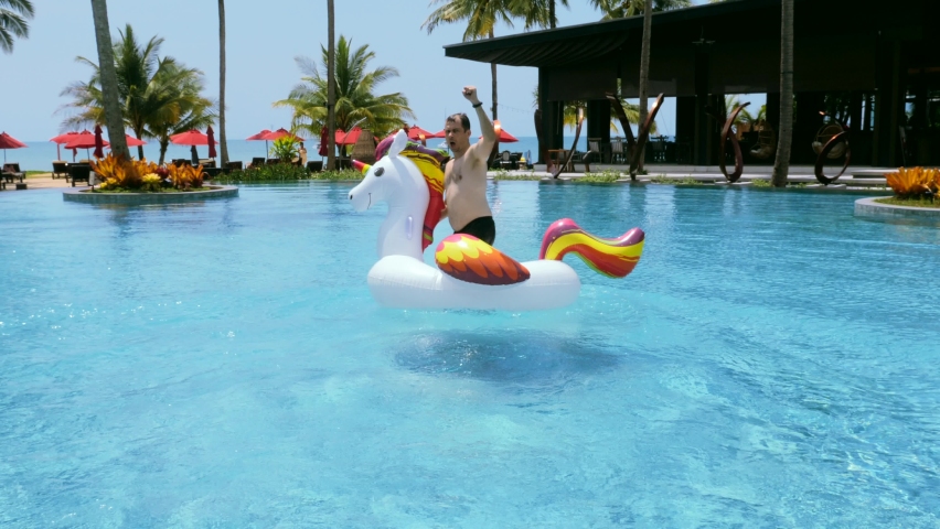 Happy adult man having fun, ride and dancing on floating inflatable unicorn in swimming pool in luxury hotel. Funny male tourist enjoying summer holidays, vacations, sunny day, sea view on | Shutterstock HD Video #1093403863
