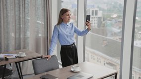 Good-looking blonde lady wearing official clothes has video-call. Woman stands near a big window communicating via video chat.
