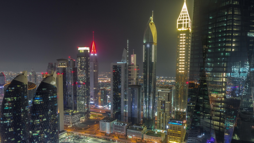 Financial center of Dubai city with illuminated luxury skyscrapers during all night timelapse, Dubai, United Arab Emirates. Aerial view with parking and towers rooftops covered by fog. Lights