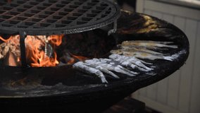 Side view of group of white floured european smelt (Osmerus eperlanus) fish frying on a barbecue on a black metal grill by open fire. Person turns over frying fish. Real time video. Selective focus.