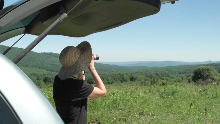 Woman solo traveler standing near car watching in binoculars a beautiful mountain view while travel driving road trip on vacation. Female enjoy sunny summer day and freedom