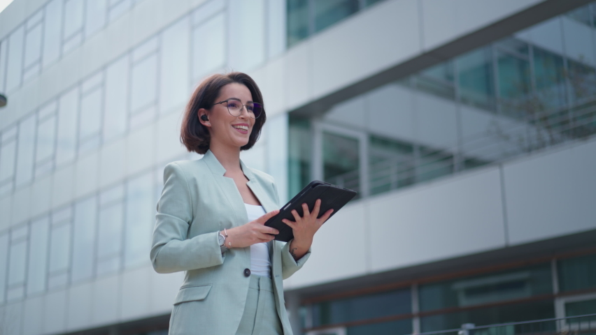 Pretty happy caucasian young woman in glasses having video chat on tablet device outdoor at business building