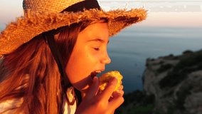 Happy girl eating corn. Summer snacking on the sea. Portrait of young beautiful woman eating grilled corn while sitting by the sea on sunset time. Close up. Slow motion.
