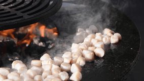 Close-up side view of scallops frying on a barbecue on a black metal grill by open fire. Real time video. Selective focus. Street food theme.