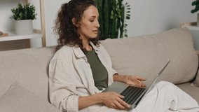 A woman with curly freelance hair sits at home on the couch, cheerfully typing on the keyboard with a smile