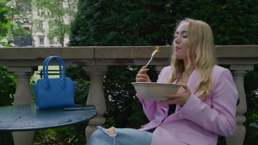 Young business Woman Girl sitting and eating in Central Park. Teen in pink jacket and blue jeans. New York City, USA. Trees on background. Lifestyle modern scene. High quality 4k footage