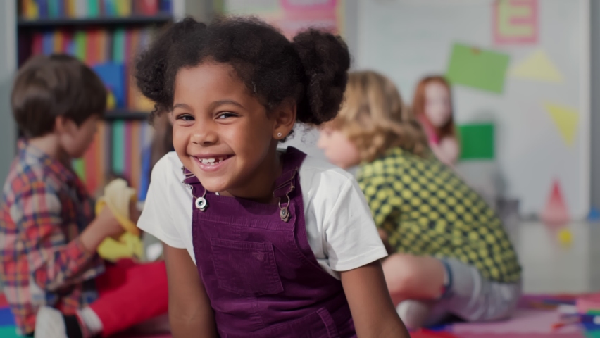 Close up portrait of smiling little African-American girl looking at camera at primary school. Adorable preschool kid sitting in playroom of kindergarten | Shutterstock HD Video #1093459677