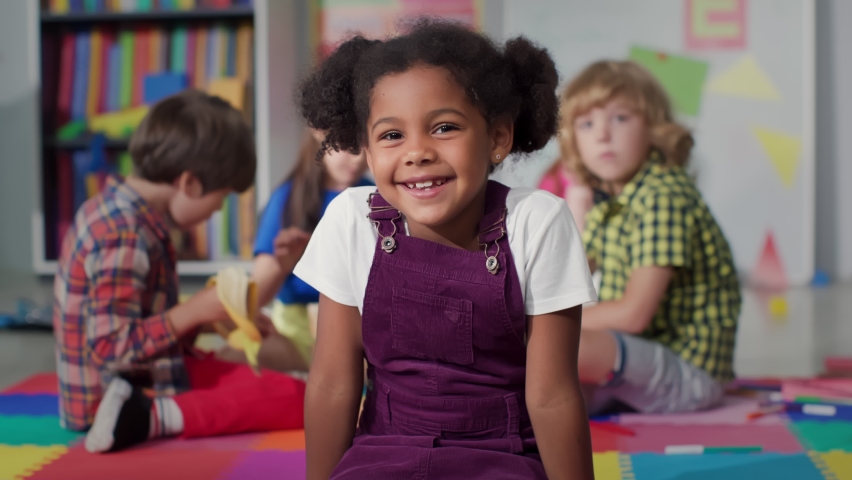 Close up portrait of smiling little African-American girl looking at camera at primary school. Adorable preschool kid sitting in playroom of kindergarten