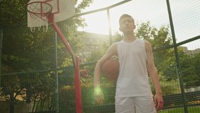 Video of a boy standing on a basketball court and holding a basketball. Cinematic shot of teenager standing with a ball in his hand and a basketball basket behind him. Basketball under his arm.
