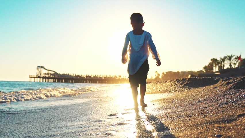 Rear view baby boy walks barefoot along seashore in water waves with foam leaving footprints on beach sand. Travel on landscape outdoor nature on summer holiday. Children walk at sun rays on sunset.
