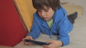 Child Playing Games In Phone at Home Lying on Couch. Boy Playing Video Game on Mobile Phone. Preschooler Plays Video Game Smartphone on Sofa. Kid Using Phone for Gaming Online Education Social Media.