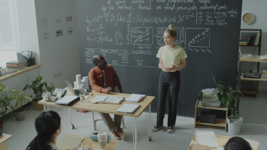 Tilt down shot of college girl standing by chalkboard in classroom and giving presentation in front of diverse group mates and African American professor Royalty-Free Stock Footage #1093467455