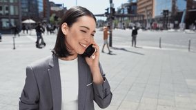 Young happy busy professional Asian business woman executive wearing suit making call on cellular holding smartphone talking on mobile phone having chat on cellphone standing on urban city street.
