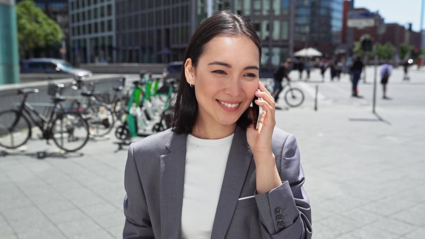 Young happy busy professional Asian business woman executive wearing suit making call on cellular holding smartphone talking on mobile phone having chat on cellphone standing on urban city street. Royalty-Free Stock Footage #1093471437
