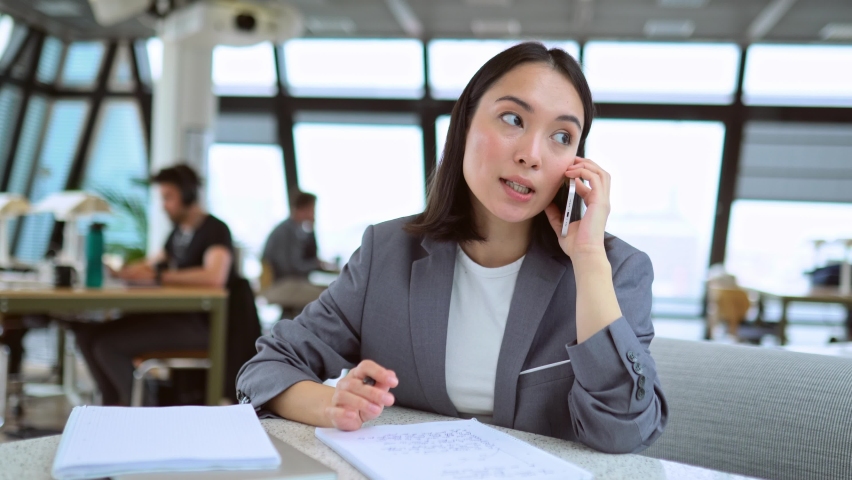 Young Chinese business woman talking on phone working in modern office. Asian businesswoman hr manager wearing suit making call on cellphone having mobile job interview sitting at work in office. Royalty-Free Stock Footage #1093471439