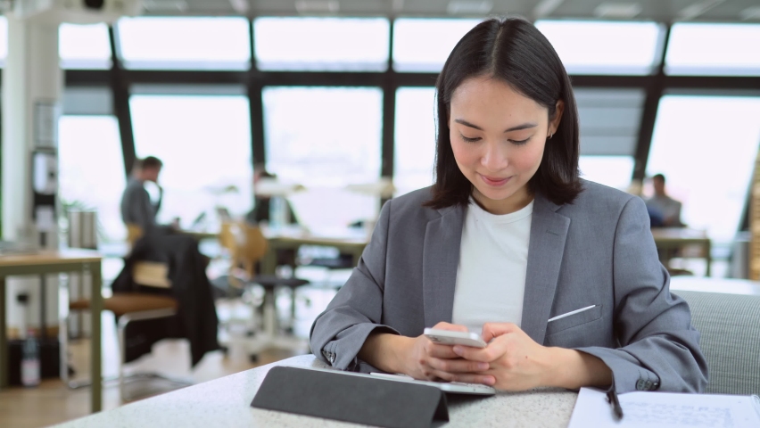 Young adult Asian business woman wearing suit working in modern office cowoking space holding smartphone. Female Chinese manager professional using mobile corporate apps on cell phone at work. Royalty-Free Stock Footage #1093471445