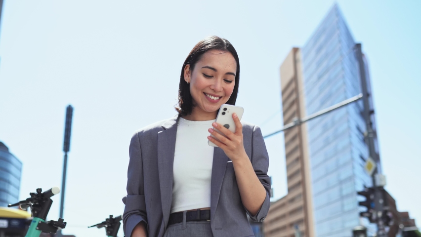 Young adult elegant successful beautiful Asian business woman, happy professional businesswoman executive holding cellphone using smartphone modern tech standing on big city urban street outside.