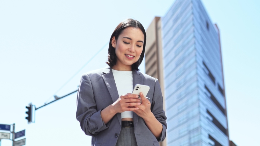 Young happy Asian woman professional executive using cell phone receiving good news in mobile message, smiling ethnic businesswoman holding smartphone looking at telephone standing on big city Royalty-Free Stock Footage #1093471451