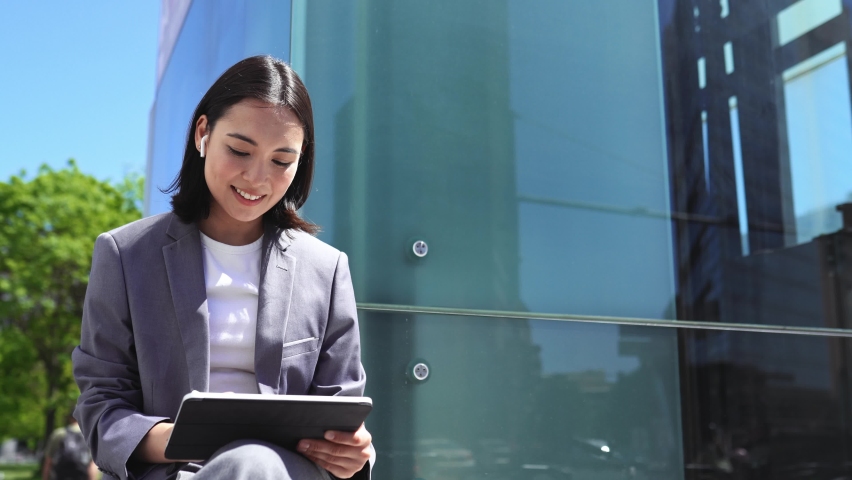 Elegant young Asian business woman executive, professional manager wearing suit holding using digital tablet modern fintech device working or e-learning online sitting in big city outside office. Royalty-Free Stock Footage #1093471455