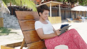 Modern Asian businesswoman working on digital tablet with internet at tropical beach in summer sunny day. Woman freelancer enjoy outdoor lifestyle work and travel on summer beach holiday vacation.