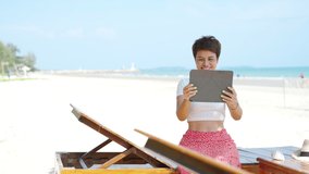 4K Modern Asian businesswoman working on digital tablet with internet at tropical beach in summer sunny day. Woman freelancer enjoy outdoor lifestyle work and travel on summer beach holiday vacation.