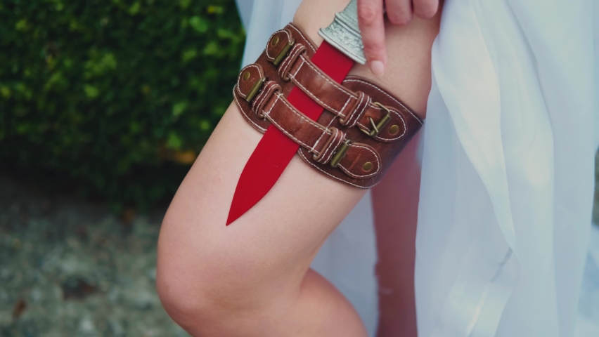 Fantasy red-haired dangerous woman medieval assassin queen with dagger in sheath on leg leather belt. White vintage old style dress. Girl princess warrior. Green summer nature trees. Hand hold knife