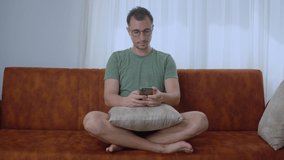 A man with a phone in the lotus position sits on a sofa and corresponds in a smartphone