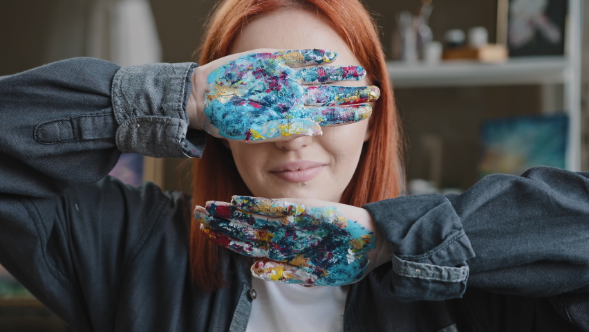 Female in art studio red-haired girl artist lady Caucasian 30s woman painter designer hides behind hands covers face with arms holding showing dirty in paint palms looking at camera smiling laugh Royalty-Free Stock Footage #1093498419