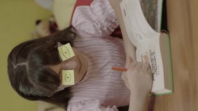 A little girl is reading a book in her room with eye-shaped papers in her glasses. Cute little girl reading a book, turning the page, learning, working, reading, reading a book.Vertical video.