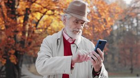 Active senior people with digital technology concept. Smiling elderly gray-haired man using smartphone for texting message,reading news,watching photo or video in social media walking in autumn park