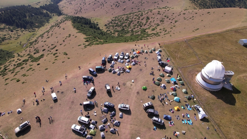 The tent camp is located near the observatory. People arrived in cars. Camp near slope. Green grass grows, in places trees. There is a large telescope with a dome. Tourists play volleyball with a