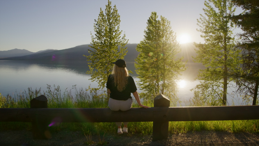 Woman enjoys pristine wilderness of the Glacier National Park. Sunset skyline reflecting in alpine lake water surface as seen from the shore. High quality 4k footage
