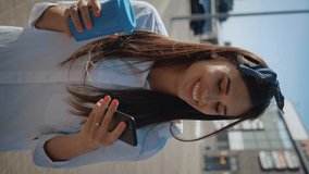 Vertical video of young woman talking on the phone and drinking coffee on background of office building. Cute smiling girl drinks coffee and uses the phone on a urban background.