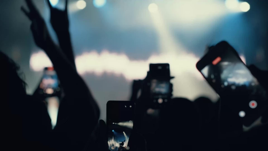 A blurry and illuminated dancing crowd. A crowd of fans standing near the stage applauding their favorite rock band. Concert in the city center. Young people with smartphones. Silhouettes of Royalty-Free Stock Footage #1093517563