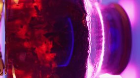 Vertical Video, Transparent Teapot with Black Tea Leaves Boils on a Gas Burner against a Neon Purple Backlight. Blurred Bubbles in Hot Water Boiling inside a Glass Kettle.