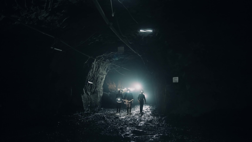 Three mining workers, in helmets, go underground through a dark tunnel. Two men and a woman. Flashlights on the helmets are on. 4K Royalty-Free Stock Footage #1093530149