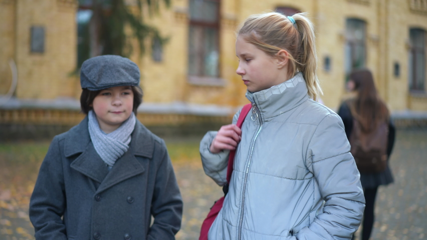 Teen girl passing backpack to boy walking with classmate on schoolyard smiling. Portrait of happy Caucasian teenage couple strolling after classes outdoors. First love and education concept Royalty-Free Stock Footage #1093536333
