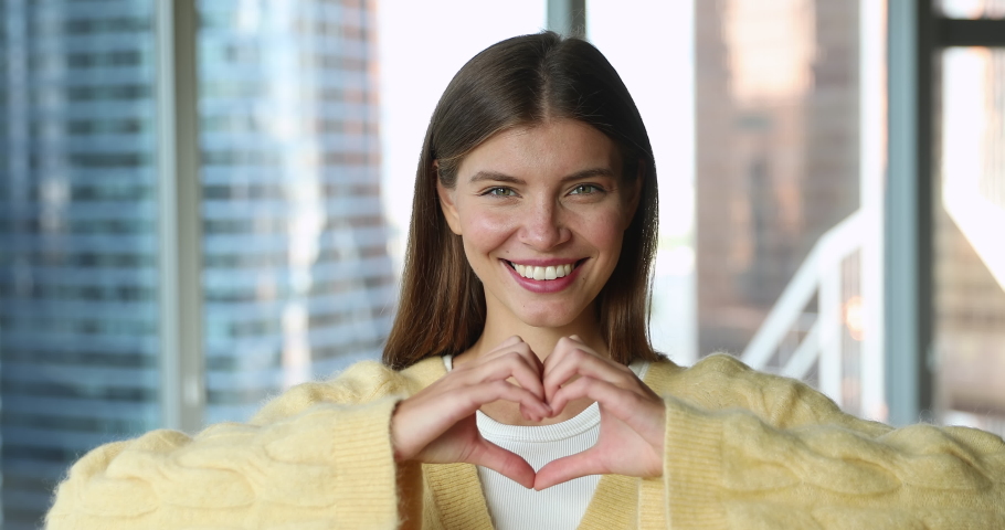 Head shot portrait smiling pretty woman in love showing heart symbol with folded fingers, pose in modern building look at camera. Affection, romantic feelings, express support, showing loyalty | Shutterstock HD Video #1093541701