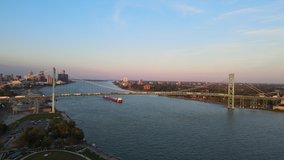 Aerial drone footage of the Ambassador Bridge crossing over the Detroit River during a beautiful summer sunset. Semi trucks crossing the bridge and a Lake Freighter passing underneath the bridge.