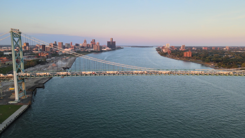 Aerial drone footage of the Ambassador Bridge crossing over the Detroit River during a beautiful summer sunset. Semi trucks crossing the bridge and a Lake Freighter passing underneath the bridge. Royalty-Free Stock Footage #1093543197