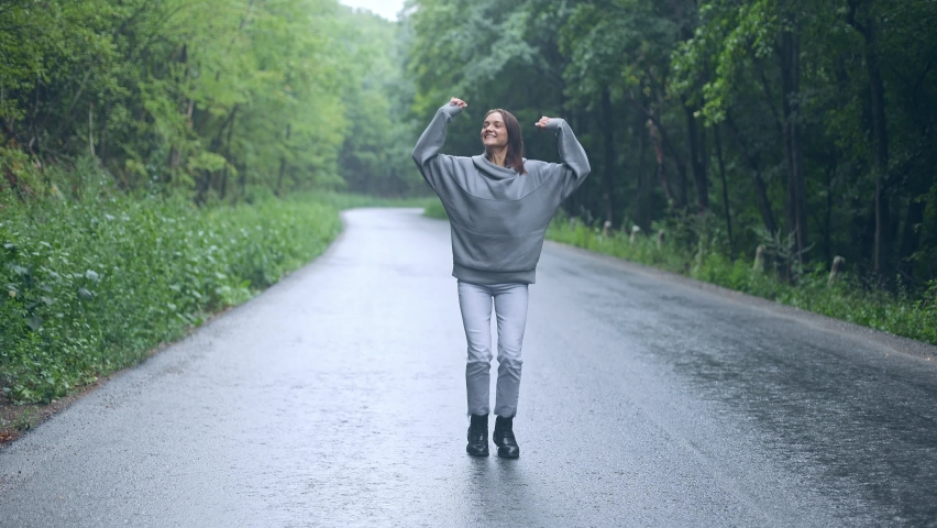 
young pretty woman girl jumping for joy dancing raising arms up, smiling, having fun, enjoying a walk, happily spending time alone, walking on a forest road in the rain