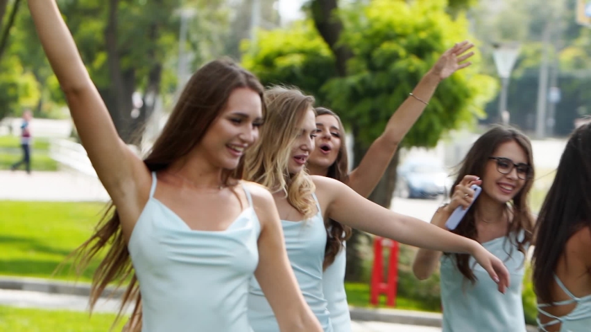 Pretty bridemaids in identical blue dresses run to hug bride together in park cheering waving hands. Woman party with friends on wedding day in elegant airy white gown and long veil. Slow motion