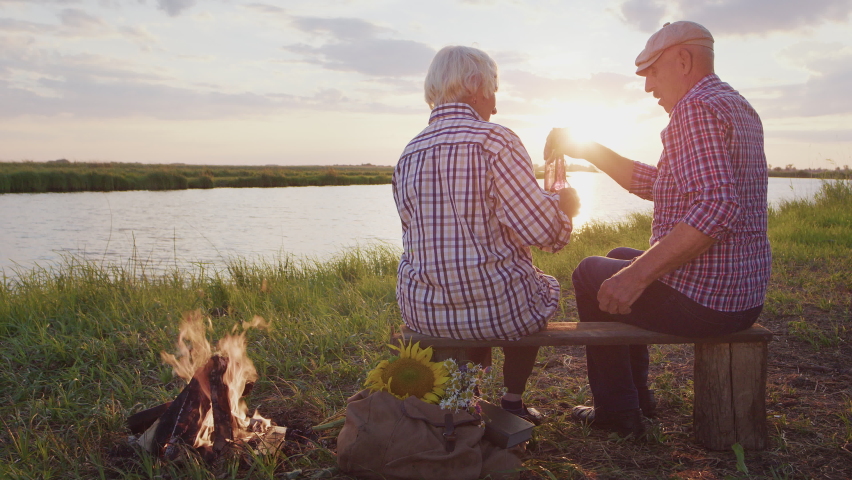 Happy old grandmother and grandfather together drink holiday beverages sitting on lake beach at sunset outdoor. Cheers anniversary celebration, friends meeting or family relax, enjoy freedom at Royalty-Free Stock Footage #1093560271