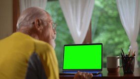 Over shoulder view of senior elderly man making video call on laptop green screen talking and listening during video chat with friends or colleagues.