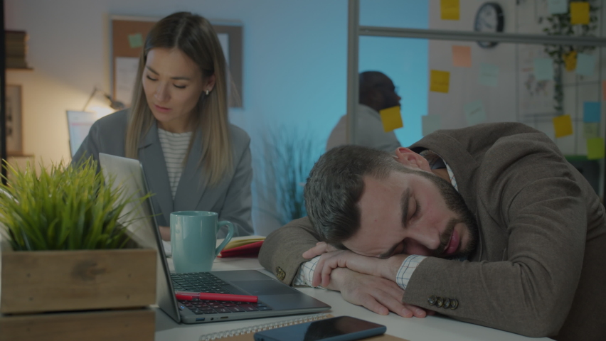 Young businessman in suit sleeping at desk while colleagues working in dark office at night. Corporate lifestyle and workplace concept. | Shutterstock HD Video #1093561351